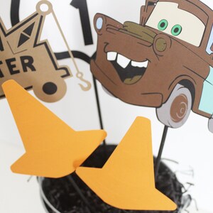 Może przedstawiać: Centralny element dekoracyjny przedstawiający brązowy, kresk&oacute;wkowy samoch&oacute;d holowniczy z uśmiechającą się twarzą i zielonymi oczami. Samoch&oacute;d otoczony jest pomarańczowymi stożkami drogowymi. Na samochodzie widnieje napis "Tow Mater".