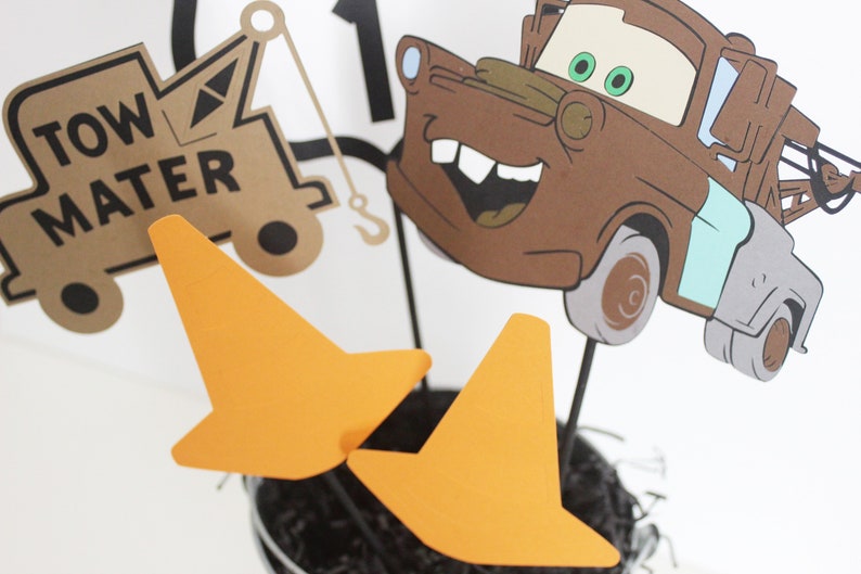 Może przedstawiać: Centralny element dekoracyjny przedstawiający brązowy, kresk&oacute;wkowy samoch&oacute;d holowniczy z uśmiechającą się twarzą i zielonymi oczami. Samoch&oacute;d otoczony jest pomarańczowymi stożkami drogowymi. Na samochodzie widnieje napis "Tow Mater".