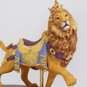 Może przedstawiać: Porcelanowa figurka lwa w koronie i kolorowym siodle, stojąca na drewnianej podstawie. Lew jest zwierzęciem karuzelowym.