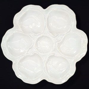 May include: White ceramic oyster plate with six individual indentations for serving oysters. The plate has a scalloped edge and a smooth, glossy finish.