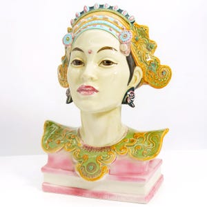 Puede incluir: Busto de cerámica de una mujer con un elaborado tocado y adornos ornamentados en los hombros. El busto presenta una tez pálida, labios rojos y ojos oscuros. El tocado está adornado con detalles azules, amarillos y rosas. Los adornos de los hombros son verdes y dorados.