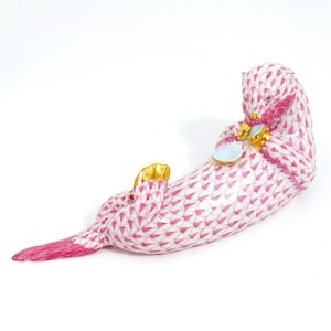 May include: A ceramic otter figurine, lying on its back. The otter is white with a pink fishnet pattern and pink accents. It has gold-coloured paws and is holding a light blue plate. The figurine is a decorative item.