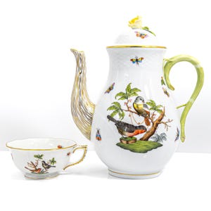 Peut inclure: Service à thé en porcelaine blanche comprenant une théière et une tasse. La théière a un bec doré et une anse verte. L'ensemble est décoré d'oiseaux colorés, de papillons et de motifs floraux. La tasse a un bord doré.
