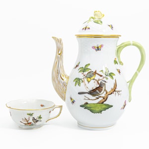 May include: A white porcelain tea set featuring a teapot and teacup. The teapot has a gold-colored handle and spout, a green handle, and a lid with a yellow rose. The set is decorated with colorful birds, butterflies, and foliage.