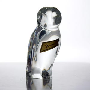 May include: A clear glass owl figurine with a gold label that reads "Baccarat". The owl is sculpted with smooth lines and a rounded head. The figurine is a decorative item.
