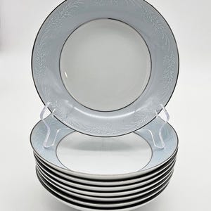 Może przedstawiać: Stosik dziesięciu białych ceramicznych misek z srebrną obwódką i delikatnym wzorem kwiatowym. Miski są ułożone na białym tle.