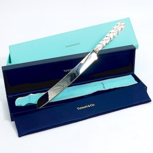 May include: A silver cake knife with a braided handle, presented in a blue box with the "Tiffany & Co." logo. The knife is displayed in a dark blue satin-lined box, with a light blue protective sleeve.