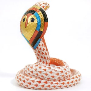 May include: A ceramic cobra figurine with a detailed, colorful design. The cobra's hood features gold, black, blue, and red patterns. The body is white with orange scale-like markings. The snake is coiled, with its head raised.