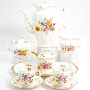 Puede incluir: Un juego de té de porcelana blanca con diseños florales. El juego incluye una cafetera, azucarero, lechera, dos tazas de té con platillos y una bandeja. Los bordes de cada pieza están adornados con detalles dorados.