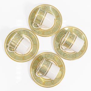 May include: Four white teacups with gold and green decorative bands and handles, each resting on a matching saucer. The saucers and cups have a gold and green ornate design. The set is arranged on a white background.