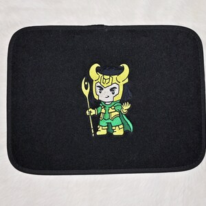 Op de afbeelding: Zwarte auto vloermat met een geborduurd beeld van een cartoonfiguur in groene en gele kleding met een staf in de hand. Het personage heeft zwart haar en een gele helm.