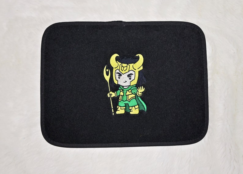 Op de afbeelding: Zwarte auto vloermat met een geborduurd beeld van een cartoonfiguur in groene en gele kleding met een staf in de hand. Het personage heeft zwart haar en een gele helm.
