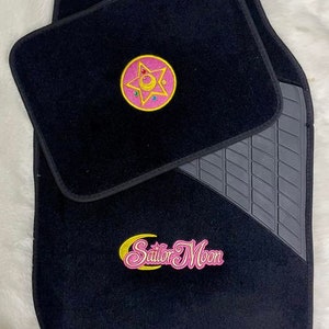 Op de afbeelding: Zwarte autovloermatten met een roze en geel Sailor Moon logo en een roze en geel sterontwerp.