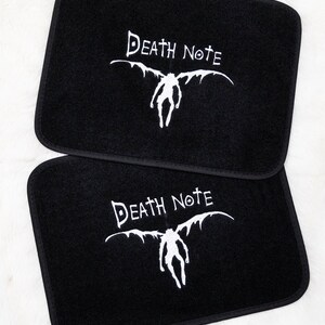 Op de afbeelding: Twee zwarte autovloermatten met een wit geborduurd Death Note-logo en het personage Ryuk met vleugels.