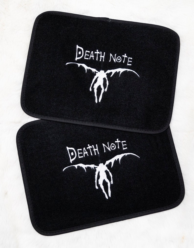 Op de afbeelding: Twee zwarte autovloermatten met een wit geborduurd Death Note-logo en het personage Ryuk met vleugels.