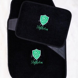 Op de afbeelding: Zwarte autovloermatten met een geborduurd Slytherin-huiswapen in groen en wit, met het woord "Slytherin".
