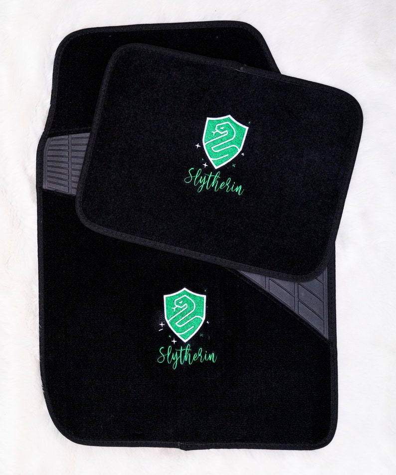 Op de afbeelding: Zwarte autovloermatten met een geborduurd Slytherin-huiswapen in groen en wit, met het woord "Slytherin".
