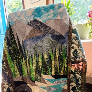 May include: A quilt with a mountain scene. The quilt is made of different fabrics and has a patchwork design. The mountains are blue, green, and brown. The sky is blue with white clouds. The quilt is hanging on a chair.