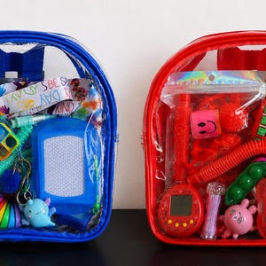 May include: Two clear backpacks, one blue and one red, each filled with a variety of colourful sensory toys and fidget items. The contents include small digital games, textured tubes, plastic figures, and patterned ribbons visible through the transparent material.