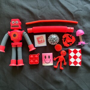 May include: A selection of red and pink fidget toys on a black background. The toys include a robot, a pop tube, a pop-it, a smiley face, a small red figure, a puzzle cube, and other sensory toys.