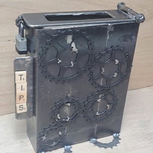 Op de afbeelding: Een donkergrijze metalen fooienbox met een gleuf aan de bovenkant. De doos is versierd met tandwielvormige uitsparingen en heeft het woord "TIPS" gespeld met lettertegels. Een hangslot beveiligt de doos.
