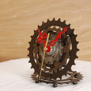 Puede incluir: Un reloj hecho con piezas de bicicleta reutilizadas. La esfera del reloj está hecha de un plato de cadena de bicicleta y las manecillas son de metal. El reloj está sobre una superficie blanca.