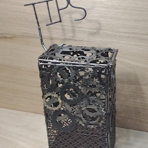 Op de afbeelding: Een metalen fooienbox met een rechthoekige vorm, gemaakt van tandwielen en metaal. Het woord "TIPS" staat bovenaan. De box heeft een donkere, industriële uitstraling.