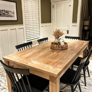 Puede incluir: Una mesa de comedor de madera con un acabado natural y sillas negras. La mesa est&aacute; puesta con una bandeja tejida, un jarr&oacute;n con flores secas y calabazas blancas.
