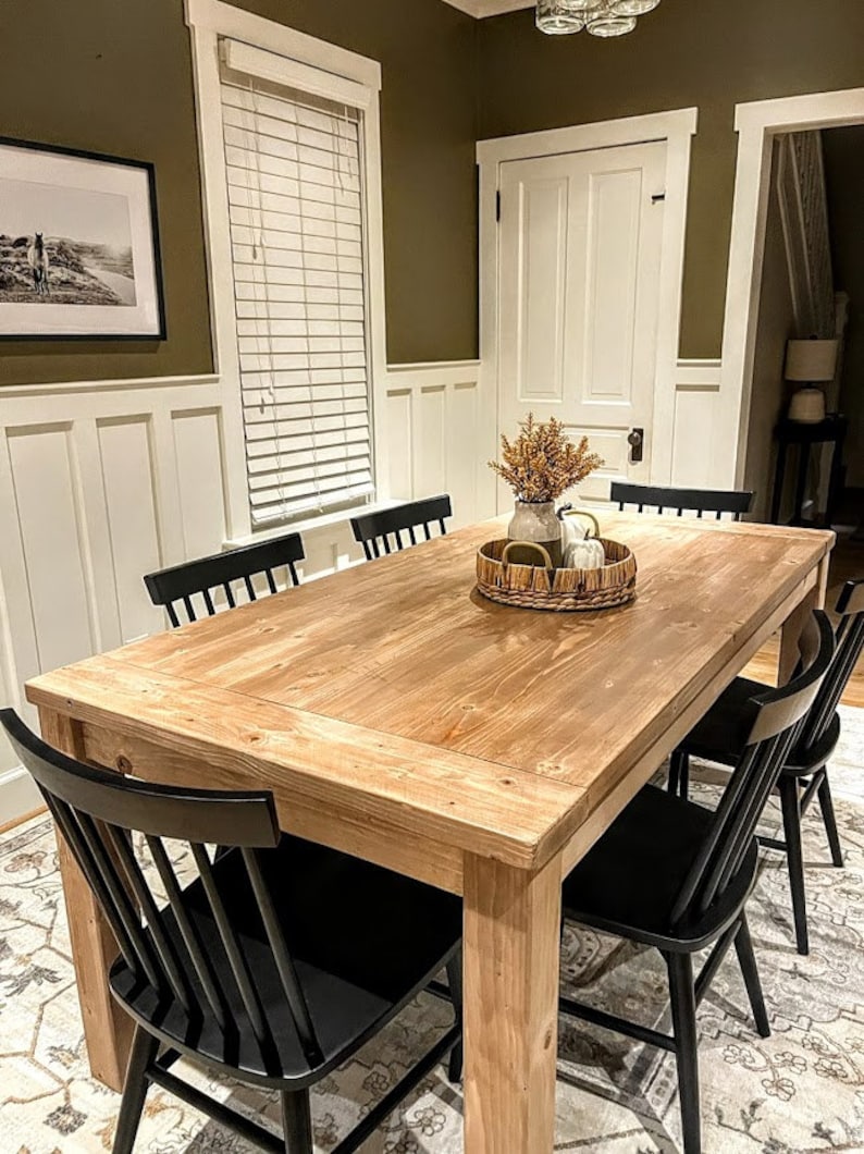 Puede incluir: Una mesa de comedor de madera con un acabado natural y sillas negras. La mesa est&aacute; puesta con una bandeja tejida, un jarr&oacute;n con flores secas y calabazas blancas.
