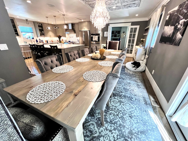 Puede incluir: Una gran mesa de comedor de madera con un acabado marr&oacute;n claro y patas pintadas de blanco. La mesa est&aacute; puesta con manteles individuales blancos y tiene ocho sillas con tapicer&iacute;a gris. Las sillas tienen respaldos capitonn&eacute;s y ribetes de tachuelas plateadas. Una alfombra gris con un patr&oacute;n floral cubre el suelo.