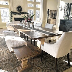 May include: A large wooden dining table with a white faux fur bench and white chairs. The table has a black and white runner and a vase of pink flowers in the center. The room has a large window with a view of a snowy landscape.