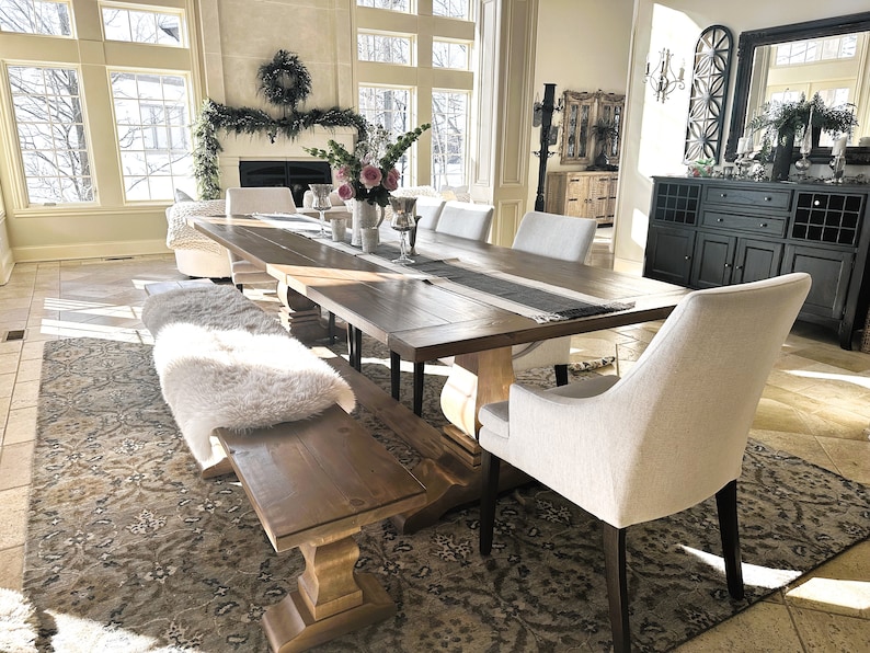 May include: A large wooden dining table with a white faux fur bench and white chairs. The table has a black and white runner and a vase of pink flowers in the center. The room has a large window with a view of a snowy landscape.