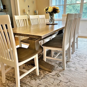 May include: A rustic wooden dining table with a natural wood finish and a pedestal base. The table is surrounded by white chairs with brown seats. The table is set with a vase of flowers and a rug with a beige and brown pattern.