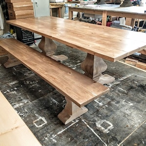 May include: A light wood farmhouse style dining table with a matching bench. The table has a rectangular top and turned legs. The bench has a rectangular top and turned legs. The table and bench are both made of unfinished wood.