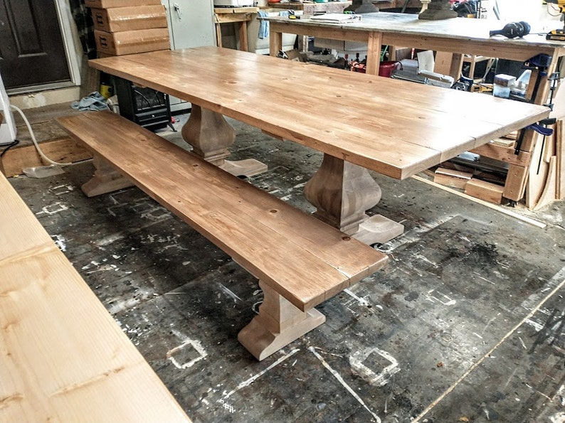 May include: A light wood farmhouse style dining table with a matching bench. The table has a rectangular top and turned legs. The bench has a rectangular top and turned legs. The table and bench are both made of unfinished wood.