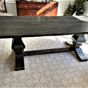 May include: A large, rectangular, dark gray wooden dining table with a trestle base. The table has a smooth, stained surface and is supported by two ornate, pedestal-style legs. The table is set on a patterned rug in a well-lit room.