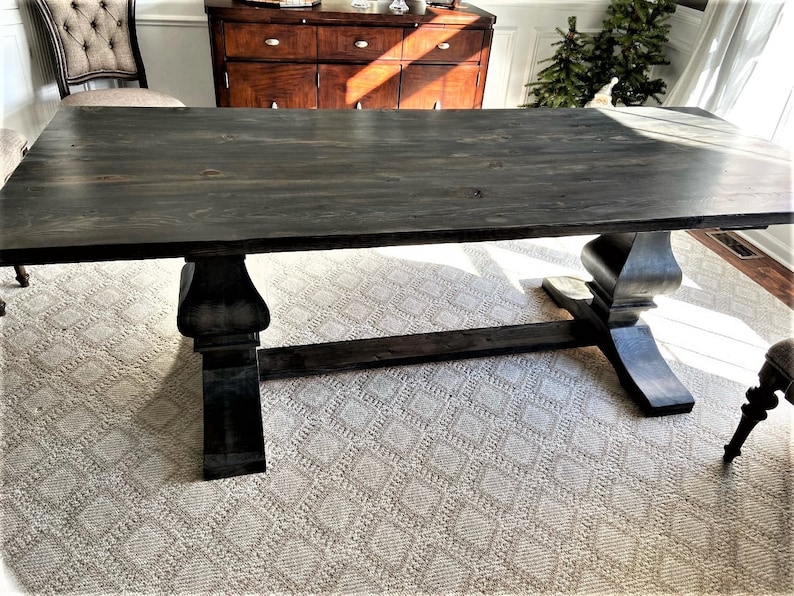 May include: A large, rectangular, dark gray wooden dining table with a trestle base. The table has a smooth, stained surface and is supported by two ornate, pedestal-style legs. The table is set on a patterned rug in a well-lit room.