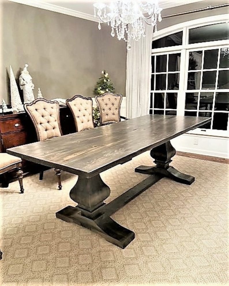 May include: A large, rectangular dining table with a dark gray, weathered wood top and a pedestal base. The table is surrounded by three upholstered chairs with button tufting. The room has a neutral color palette and a large window.
