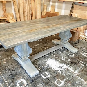 May include: A large, rectangular wooden dining table with a gray-washed finish. The table features a pedestal base with ornate, turned supports. The tabletop shows the natural wood grain. The table is a rustic, farmhouse style.