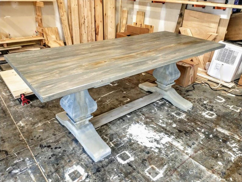May include: A large, rectangular wooden dining table with a gray-washed finish. The table features a pedestal base with ornate, turned supports. The tabletop shows the natural wood grain. The table is a rustic, farmhouse style.