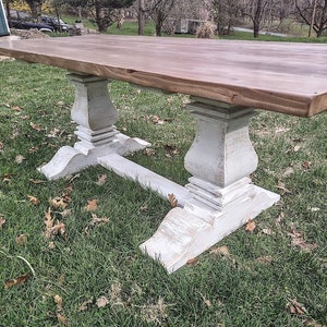 May include: A large, rectangular wooden table with a natural wood top and distressed white painted base. The table features a classic pedestal design with ornate detailing and is set on a grassy lawn.