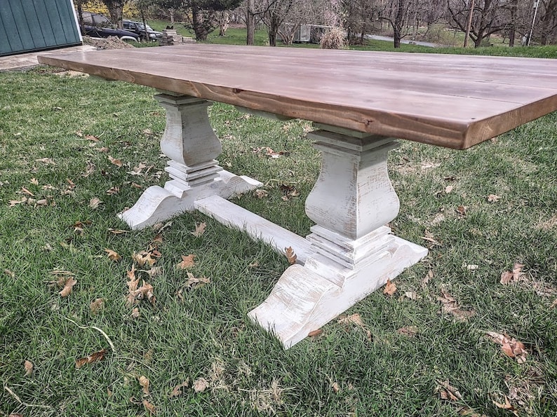 May include: A large, rectangular wooden table with a natural wood top and distressed white painted base. The table features a classic pedestal design with ornate detailing and is set on a grassy lawn.