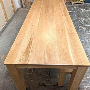 May include: A large, rectangular wooden table with a light brown finish. The table has a solid wood top and four sturdy, square legs. The wood grain is visible on the tabletop and legs.