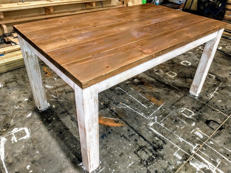 Puede incluir: Una mesa de madera r&uacute;stica con una base blanqueada. La superficie de la mesa est&aacute; hecha de tablones de madera natural y tiene un acabado te&ntilde;ido marr&oacute;n. Las patas de la mesa son blancas con un acabado desgastado.