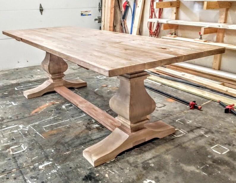 May include: A large, unfinished wooden table with a rectangular top and a pedestal base. The table is made of light-colored wood and has a natural finish. The base of the table is made of four legs that are connected by a stretcher.