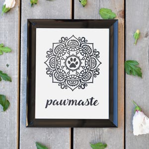 May include: Black and white print with a mandala design featuring a paw print in the centre. The print is framed in a black frame and has the text "pawmaste" below the mandala.