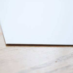 Puede incluir: Primer plano del borde de una hoja de papel blanca sobre una superficie de madera marr&oacute;n claro.