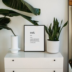 May include: A black framed print with the word "cat" in bold white letters. The definition of the word "cat" is written in smaller white letters below the word "cat". The print is sitting on a white dresser with two green plants in white pots.