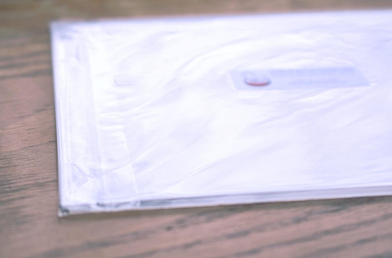 Puede incluir: Una bolsa de pl&aacute;stico blanca con una etiqueta roja y blanca. La bolsa est&aacute; doblada y apoyada sobre una superficie de madera.