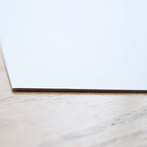 May include: A close-up of the edge of a white sheet of paper on a light brown wooden surface.